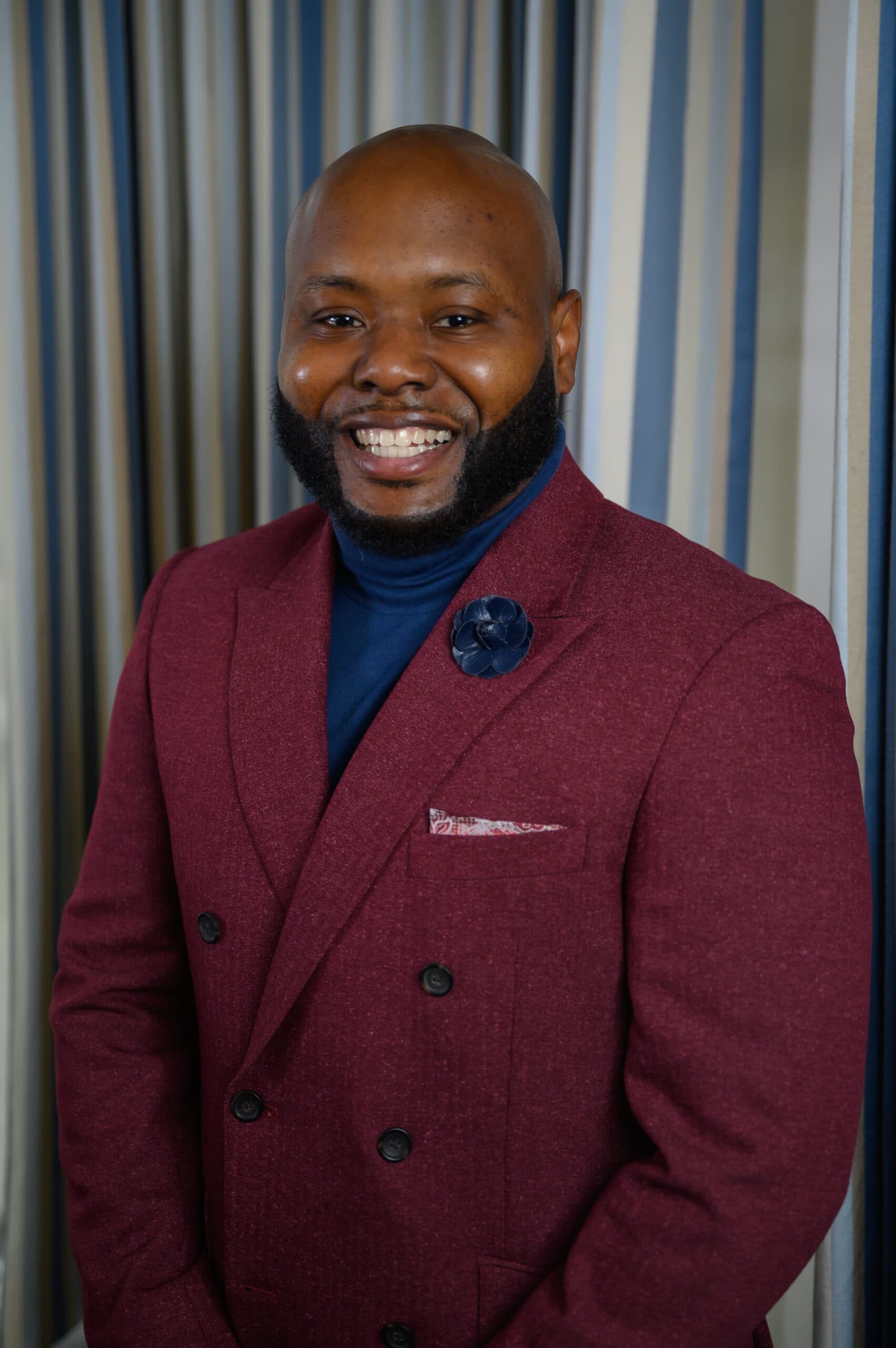 Jarel Jackson stands in front of a gray backdrop. He is wearing a maroon blazer with a navy turtleneck underneath.