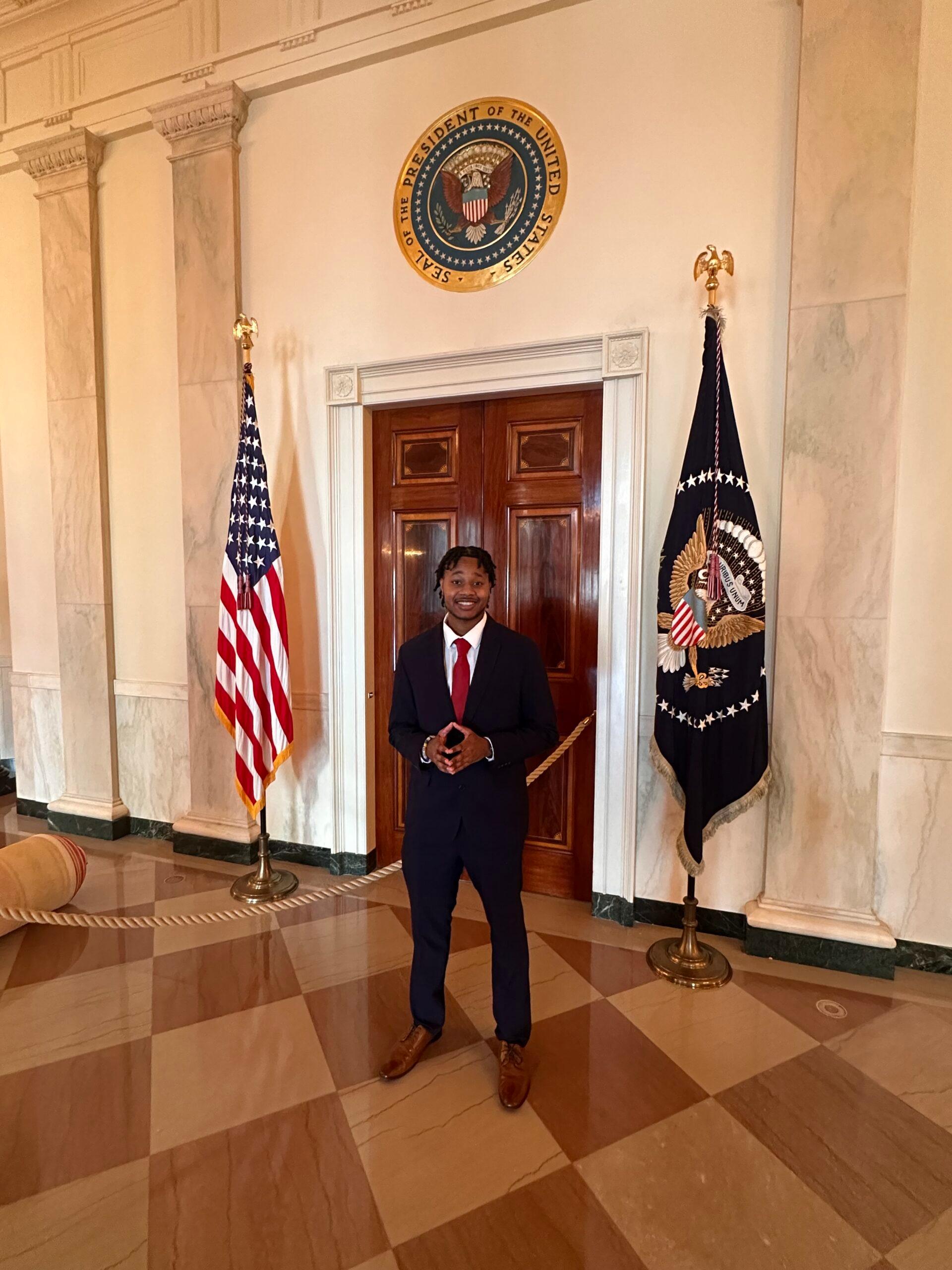 A male student stands in front of a closed door inside the Capitol building, with the United States flag and the Great Seal of the United States flag hanging from standing poles on either side of him.