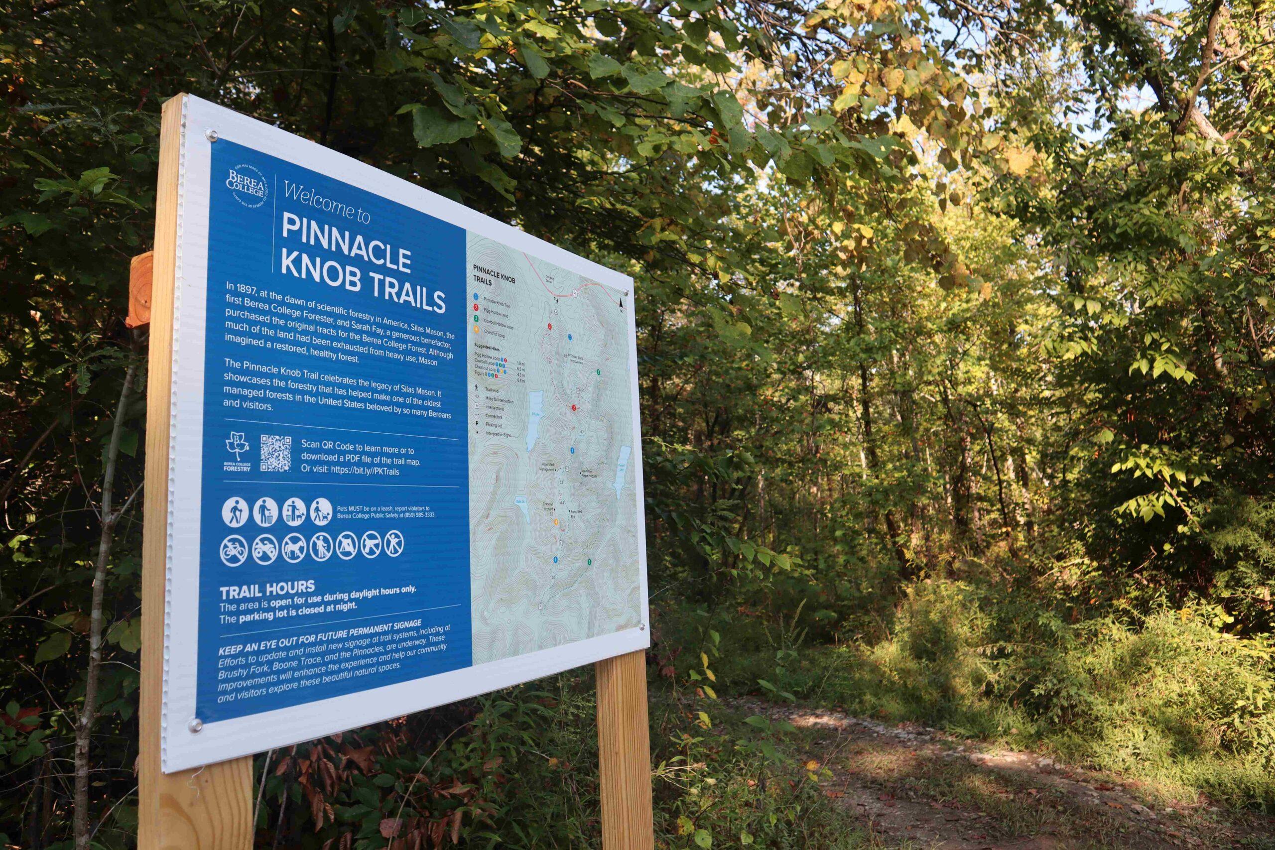 A sign introducing the new Pinnacle Knob Trails is shown near the left side of the frame, with sunlight shining on trees in the background.